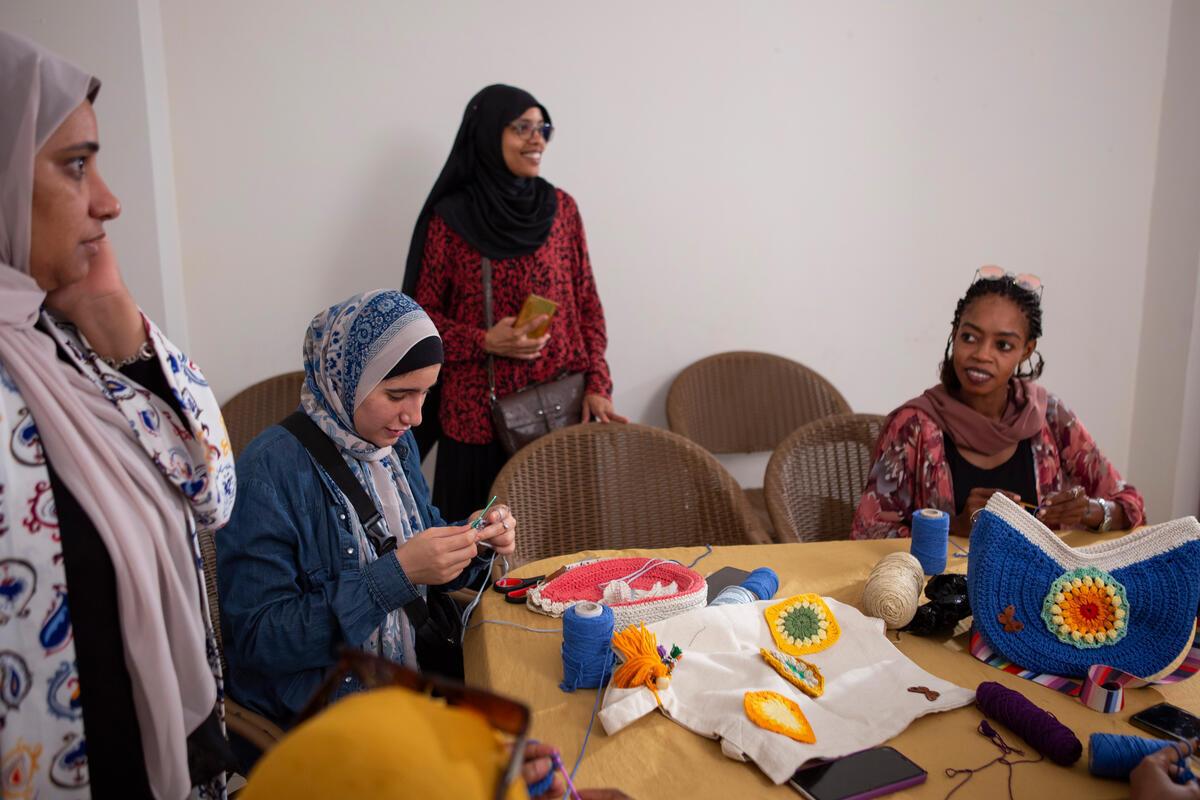 Two women sit at a table crocheting while two other women stand.